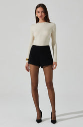 A woman is wearing a cream-colored, long-sleeved, ribbed knit sweater with a round neckline. She is also wearing high-waisted black shorts and sheer black tights. Her outfit is accessorized with gold hoop earrings, a gold bracelet on her right wrist, and a gold ring on her right hand. She has black pointed-toe high heels.